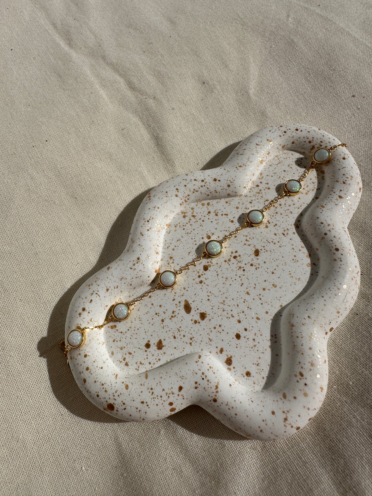 White ceramic dish with speckled texture holding a delicate gold opal bracelet on a beige fabric background