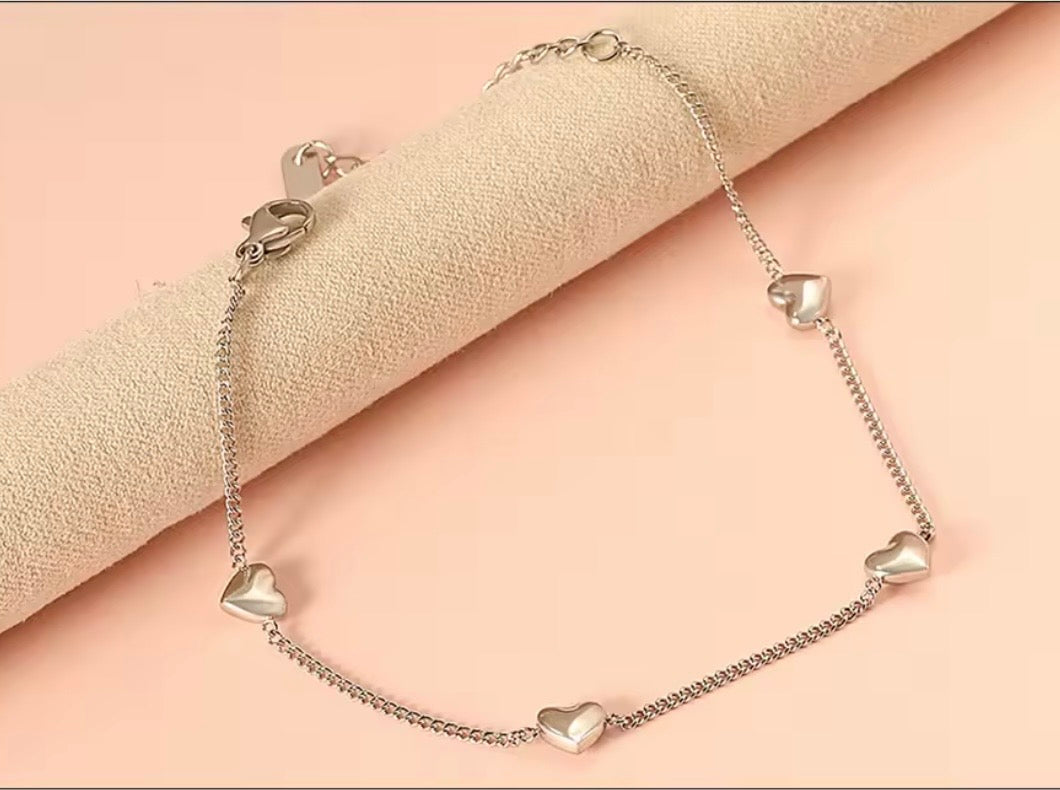A silver chain bracelet  with five small heart charms, displayed on a beige textured cylindrical stand against a soft peach-pink background.