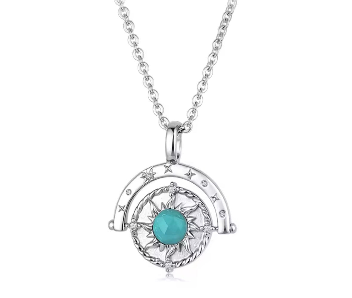 A silver-toned necklace featuring a spinner celestial pendant with a central turquoise-blue faceted gemstone and small clear stones, shown on a plain white background.