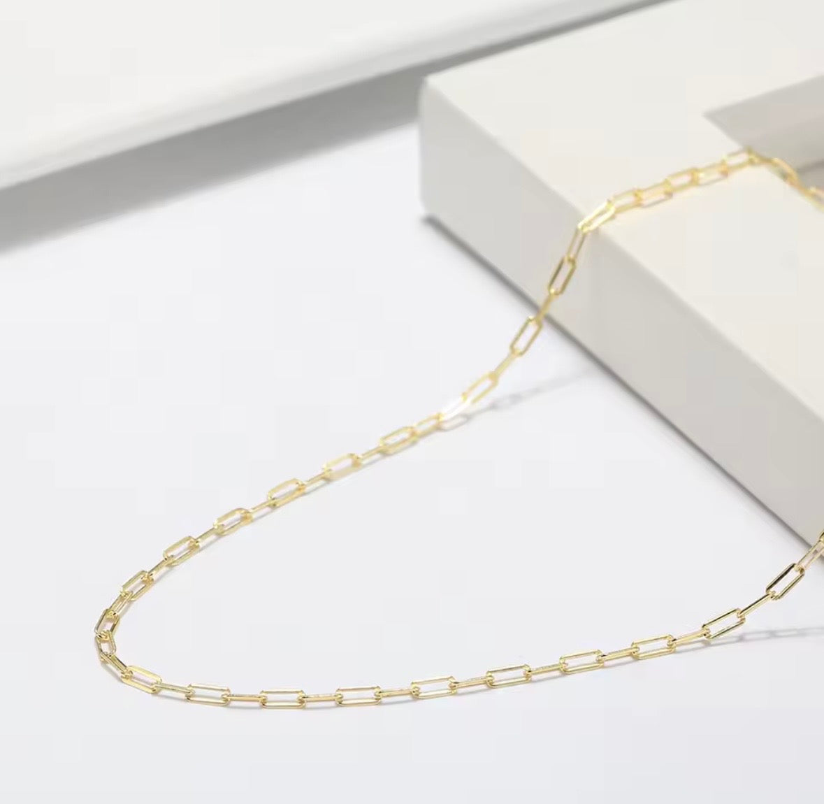 Gold fine paperclip chain necklace on a white surface with a white box in the background